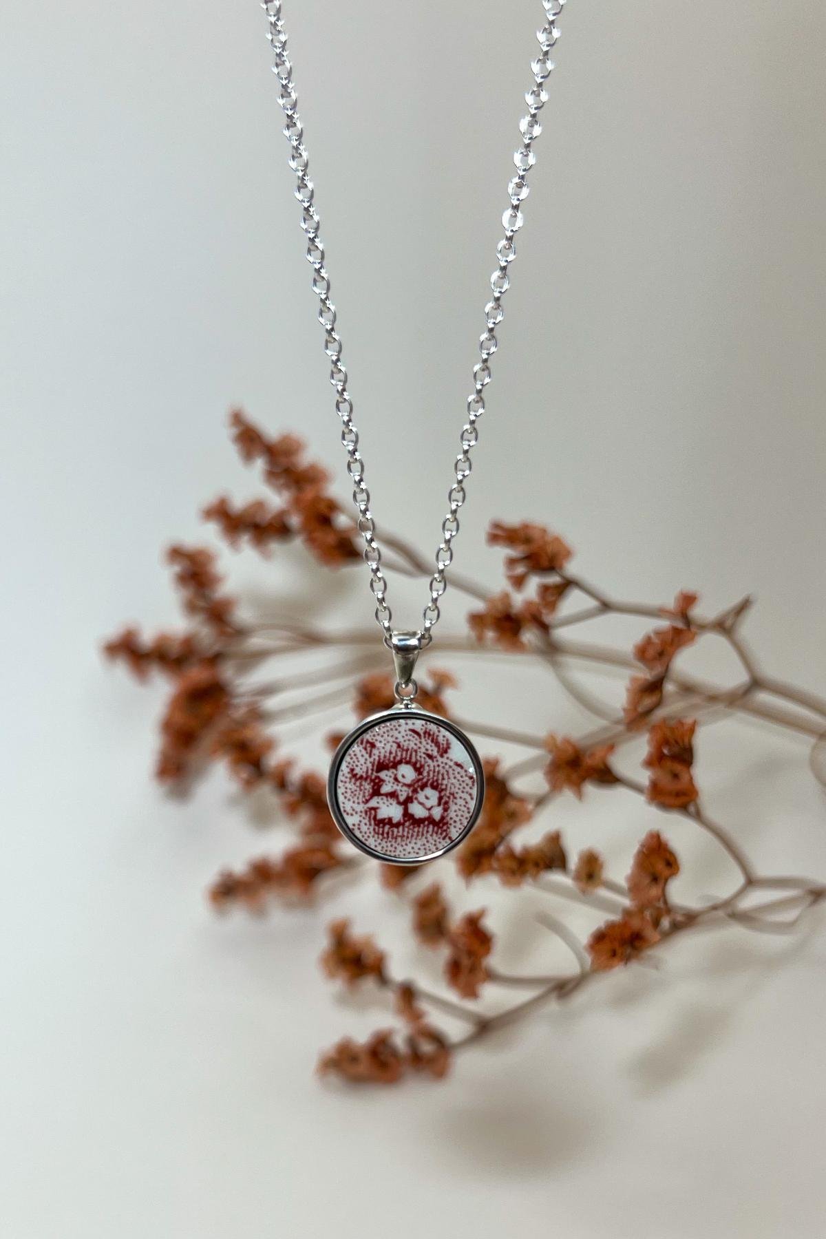 Pendentif en faïence ancienne rouge à motif floral monté sur une chaîne en argent, présenté sur un fond neutre avec une branche séchée. Bijou fait main signé Encore Beaux.